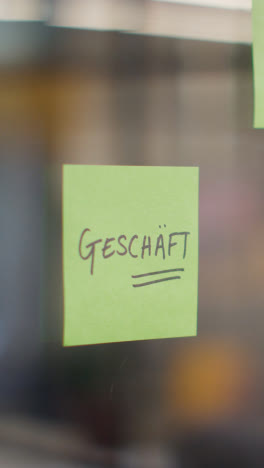 Vertical-Video-Close-Up-Of-Woman-Putting-Sticky-Note-With-German-Word-Geschaft-Or-Shop-Written-On-It-Onto-Transparent-Screen-In-Office-